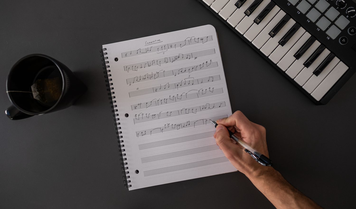 Music composition notebook on gray background alongside keyboard with hand writing 